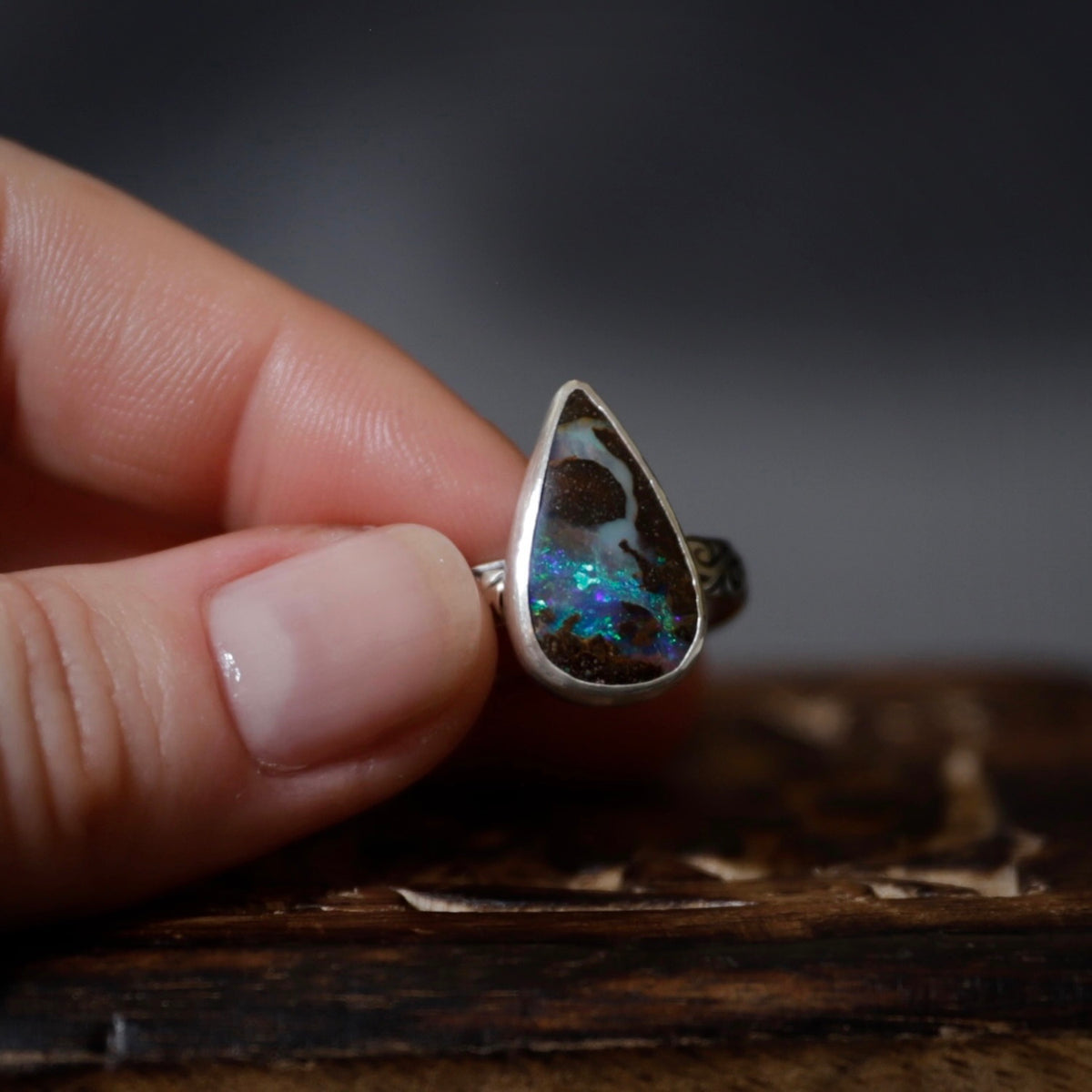 A hand holding a silver ring with a teardrop-shaped opal gemstone that displays vibrant blue, green, and orange flashes against a dark, blurred background.