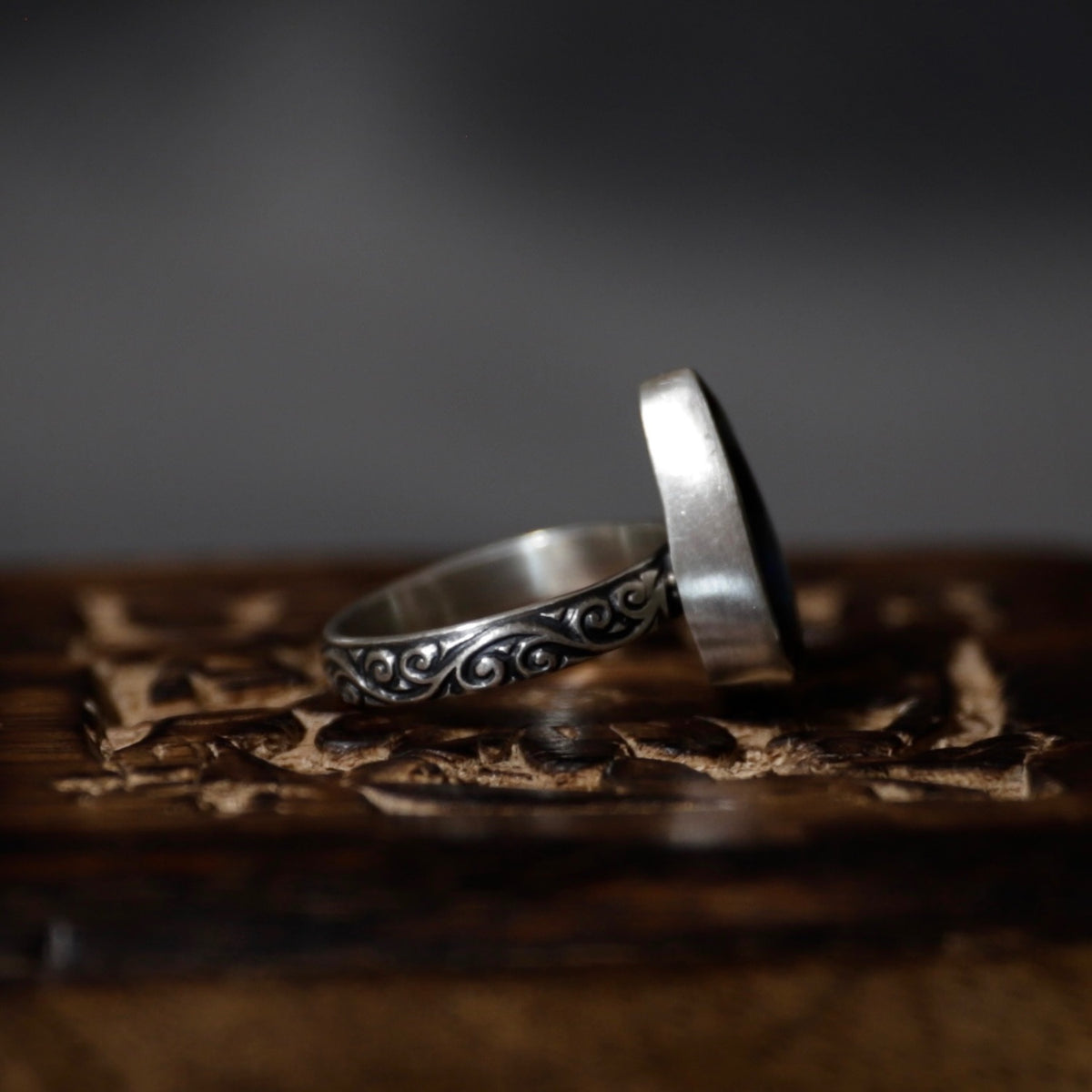 A silver ring with an engraved scrollwork pattern, shown standing on a carved wooden surface against a dark, moody background.