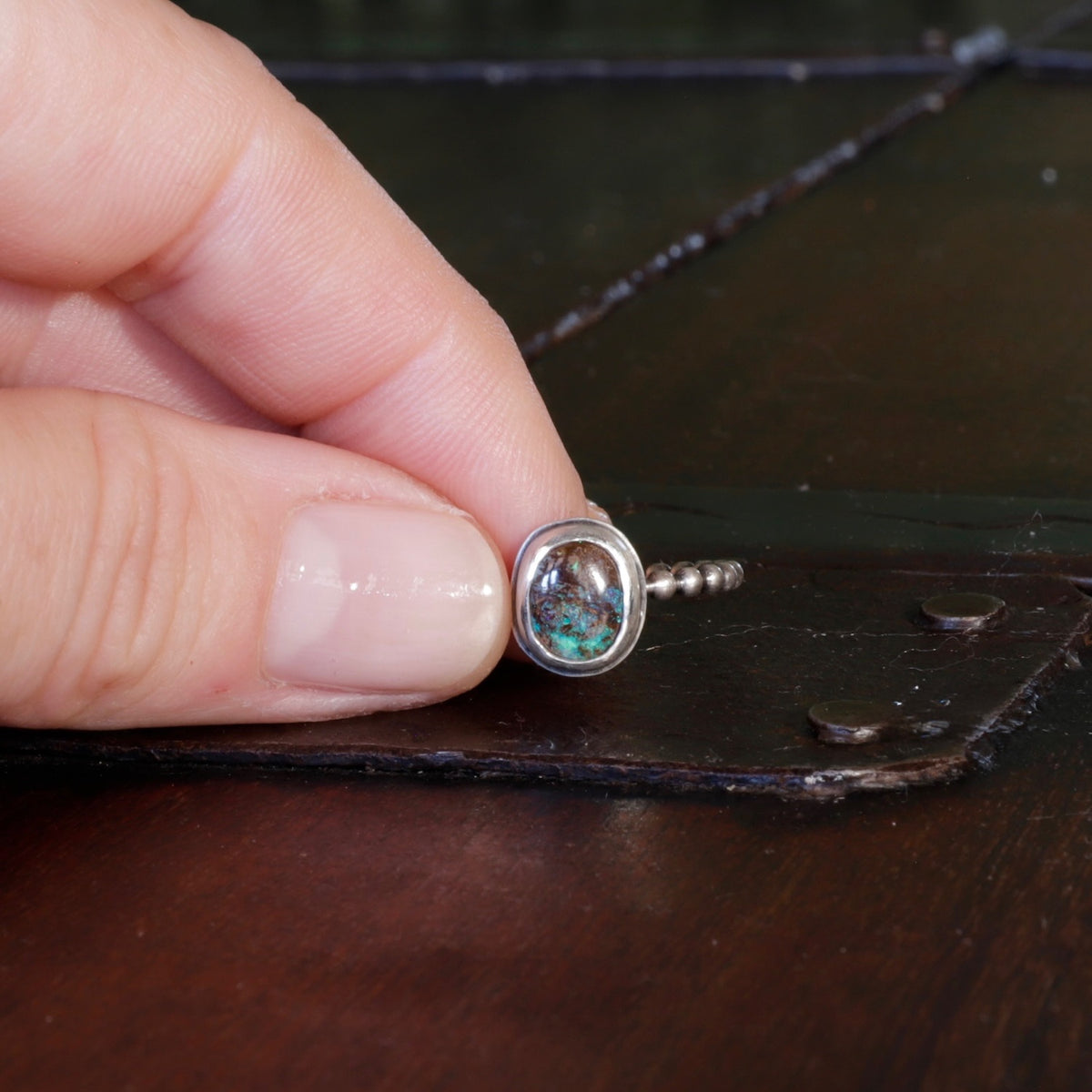 A close-up of a hand holding a small ring with a round boulder opal cabochon stone that has blue, green inclusions, resting on a dark brown surface.