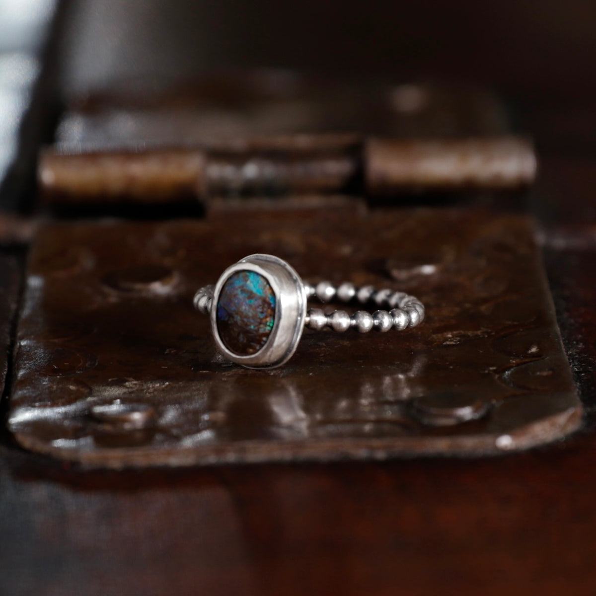 A silver beaded ring with a round bezel-set Australian boulder opal gemstone, resting on a rustic, textured dark metal surface.