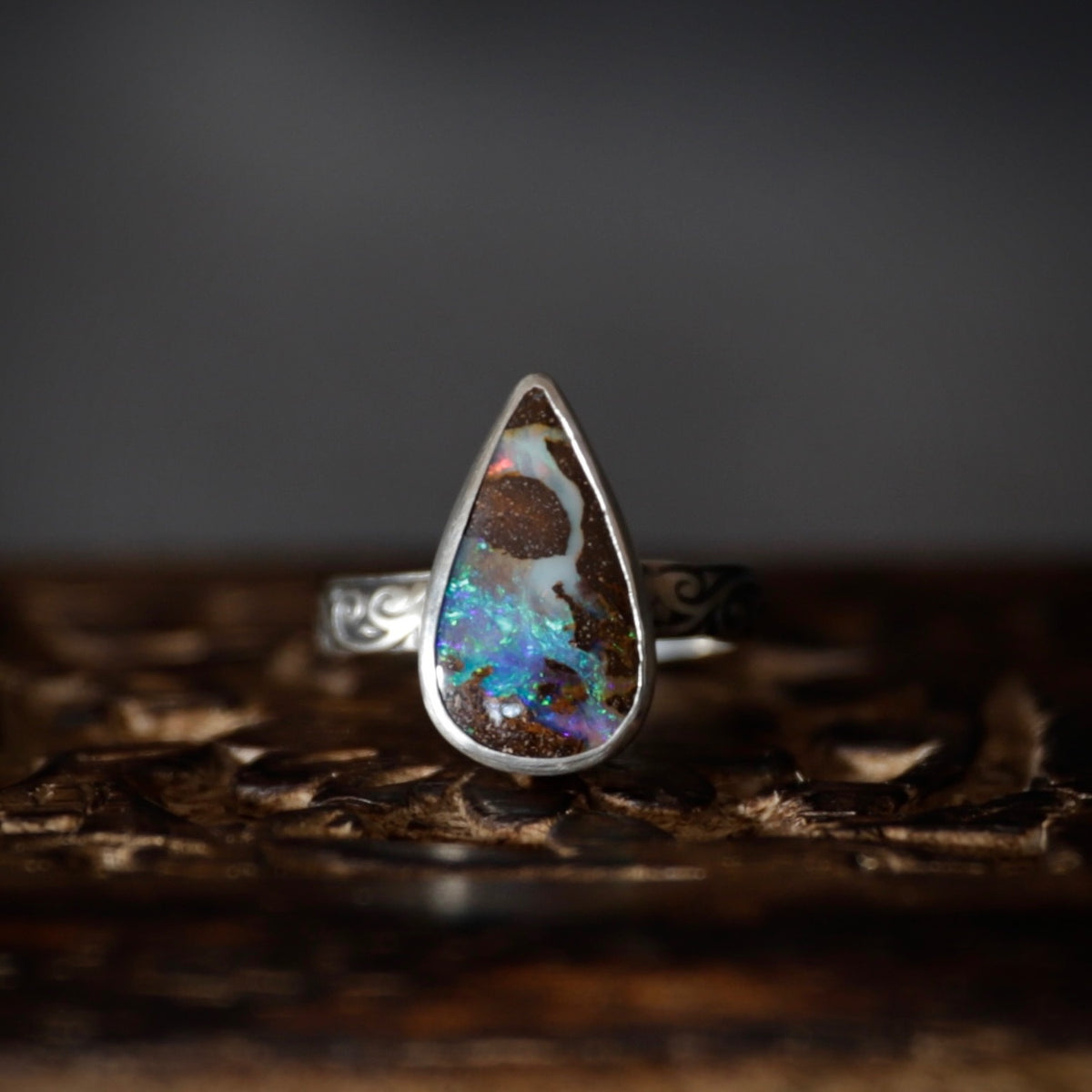 A silver ring featuring a teardrop-shaped opal gemstone with vibrant blue, green, and orange flashes against a dark brown matrix, displayed on a carved wooden surface.