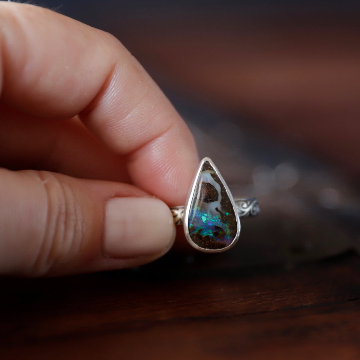 A close-up of a hand holding a silver ring with a teardrop-shaped blonder opal gemstone that displays vibrant blue, green flashes against a dark wooden background.