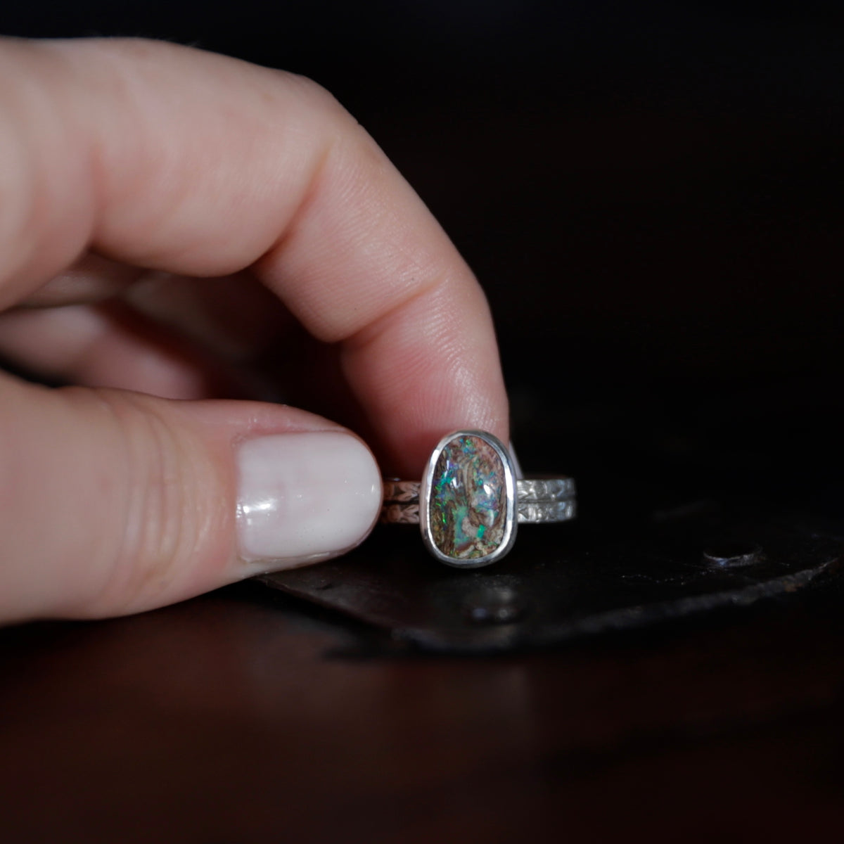 A hand holds a silver ring featuring a oval-shaped wood opal gemstone. The ring is photographed against a dark, out-of-focus background.
